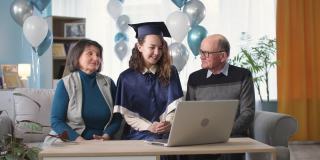 在线毕业典礼，快乐的父母和女毕业生高兴地鼓掌在毕业典礼上通过视频连接，而坐在家里的沙发上，背景是气球
