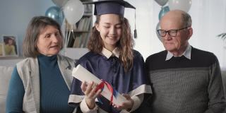 肖像幸福的家庭和研究生的女儿，戴着学位帽，穿着学位服，手里拿着文凭，在学年结束时，在远程教育