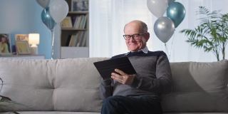 在线派对，快乐的祖父坐在客厅以气球为背景的沙发上，在社交网络上发短信，庆祝假期