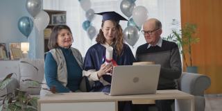 在线毕业，快乐的年轻女子在衣帽和她的父母在一个在线典礼上通过笔记本电脑视频连接庆祝学年结束
