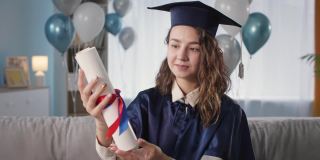 快乐的毕业生肖像，穿着学术袍和帽子，手里拿着文凭，快乐地从大学毕业，坐在沙发背景的气球在大流行期间