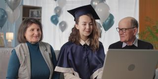 毕业证书在线，愉快的毕业生戴着帽子和长袍与父母一起庆祝大学毕业典礼期间通过视频连接坐在家里的笔记本电脑背景气球