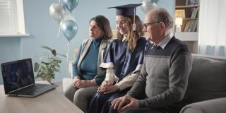快乐的大学毕业生与父母在毕业典礼上开心的拍手在线坐在家里的客厅，远程学习