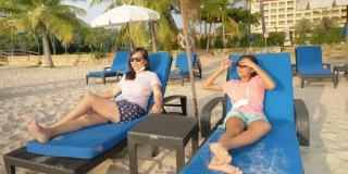 快乐的亚洲女孩戴着太阳镜和她的母亲一起放松在沙滩椅在酒店或度假村度假，与家庭，生活理念。