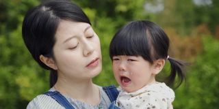 特写镜头悲伤的日本小女孩哭着指着远处，而她慈爱的母亲在公园里抱着她，拍着她的胸部安慰她
