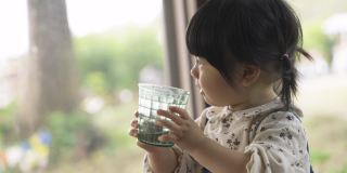 在一家餐馆里，一个天真的亚洲小女孩举起吐司，从窗边的杯子里喝水，做出滑稽的表情