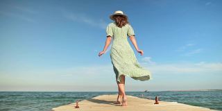 夏天，一位身穿绿色连衣裙、头戴草帽、身材苗条、年轻迷人的白人女子沿着海边的木码头散步。在假期和旅行中放松和放松