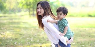 年轻的亚洲母亲背着可爱的小男孩在公园里玩耍。妈妈让婴儿背在背上。幸福的家庭和友谊的概念。