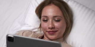 美丽的金发女人浏览平板电脑社交媒体微笑，而躺在白色的床上
