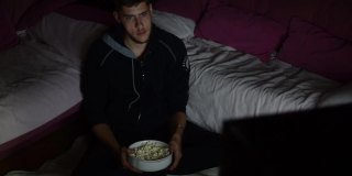 一个青少年坐在家里的地板上，在黑暗中看电影，吃爆米花，喝汽水，享受他的电影之夜。