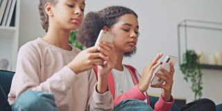 两个非裔美国女孩坐在沙发上玩智能手机，沉迷于电子产品
