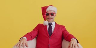 年轻的八字胡电视推销员穿着红色的衣服，戴着圣诞老人的帽子，戴着红色滑稽的眼镜，坐在椅子上，突然收到了一个惊喜的礼物并说话。新年销售和促销惊喜哇概念