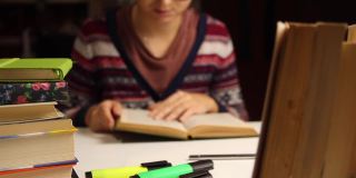模糊的年轻女子戴着眼镜在晚上研究，思考。千禧学子读书。书和记号笔放在前景上。教育学习观念