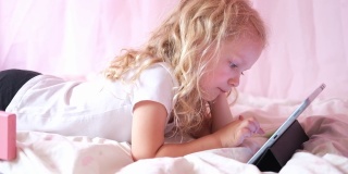 高加索好奇卷曲可爱的学龄前女孩使用数字平板技术设备躺在床上独自。小孩子拿着垫子在家里上网玩游戏。儿童科技上瘾