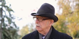 一位戴着眼镜和黑帽子的老爷爷在秋天的城市公园里散步
