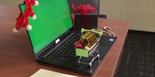 在绿色屏幕和圣诞装饰背景的笔记本电脑键盘上，年轻女性的手拿着装满小礼物的购物车。交付的概念。模拟的内容