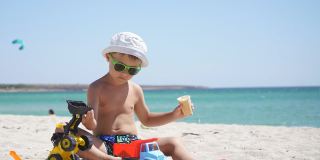 一个阳光明媚的夏日，一个男孩在沙滩上玩沙子和玩具、汽车。一个孩子坐在以蔚蓝大海为背景的沙滩上吃冰淇淋