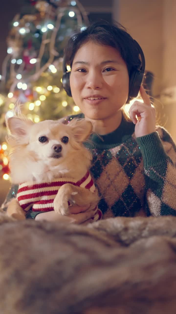 亚洲女性穿冬季布手握哈巴狗与毛衣圣诞服装享受交谈照片自拍智能手机与幸福微笑与圣诞树和灯bokeh pov垂直镜头