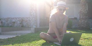 一位女士坐在阳光明媚的公园的草坪上使用她的笔记本电脑。一个白人女人坐在公园里用笔记本电脑工作。在公园里远程工作的自由职业者带着笔记本电脑坐在草地上