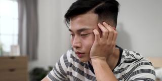 一个疲惫的中国年轻人抱着脸，努力不睡着，同时在他的卧室在互联网上学习封锁期间。