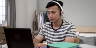 在封锁期间，戴着耳机的亚洲年轻男性在卧室里用笔记本电脑学习时，微笑着看着摄像头