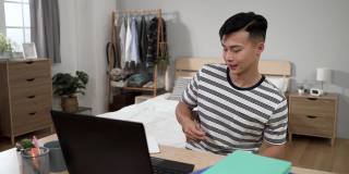一个亚洲大学生在桌子上敲着鼓，用笔按着笔记本电脑键盘上的键，满心欢喜地做完了家里的功课。