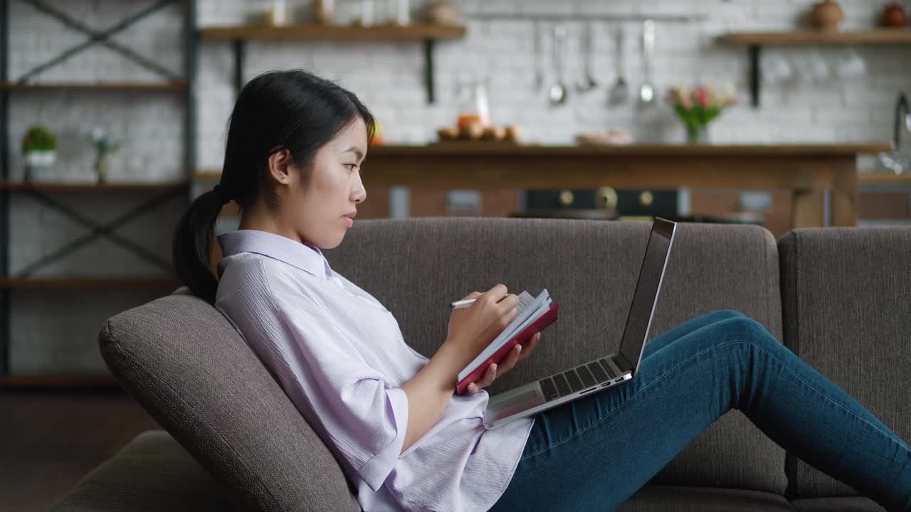 亚洲妇女使用笔记本电脑远程学习，在舒适的家里躺在沙发上写笔记。女学生在家里的沙发上用电脑进行视频通话和观看在线课程