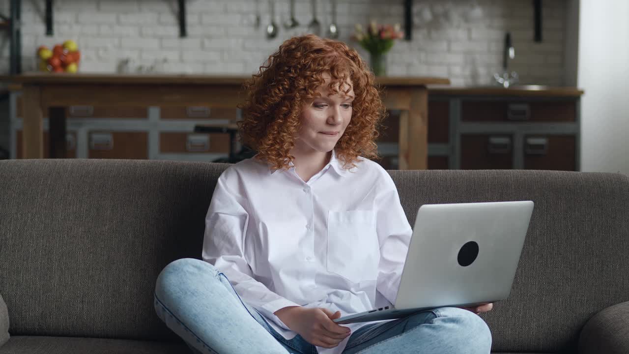 快乐的红发卷发女人用笔记本电脑聊天，坐在家里的沙发上通过在线视频聊天。