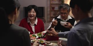 选择焦点与越过肩膀看微笑的亚洲老夫妇的祖父和祖母在新年前夕的餐桌上拿着食物和给他们的儿媳