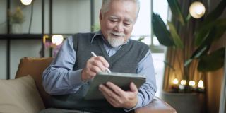 亚洲老人退休老人手使用平板电脑冲浪社交媒体浏览朋友在线社交交流距离连接，幸福亚洲老人呆在家里使用平板电脑在家里