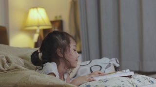 可爱的5岁亚洲女孩正在看书，躺在床上，有一盏灯发出美丽的黄色灯光。它是儿童学习、放松和发展创造力的睡前故事。视频素材模板下载