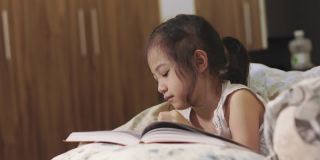 可爱的5岁亚洲女孩正在看书，躺在床上，有一盏灯发出美丽的黄色灯光。它是儿童学习、放松和发展创造力的睡前故事。