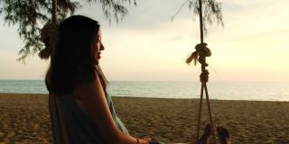 亚洲美女坐在海边的秋千上与夕阳背景在周末假期。快乐独自旅行的概念。4 k慢动作。