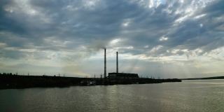 傍晚在河上的工业区域。带有烟囱的大型工厂在多云的天空下释放出黑色的烟雾。工业对河流的污染。
