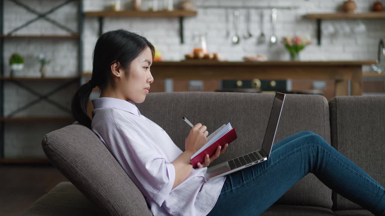 亚洲妇女使用笔记本电脑远程学习，在舒适的家里躺在沙发上写笔记。女学生在家里的沙发上用电脑进行视频通话和观看在线课程