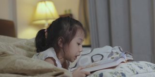 可爱的5岁亚洲女孩正在看书，躺在床上，有一盏灯发出美丽的黄色灯光。它是儿童学习、放松和发展创造力的睡前故事。