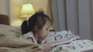 可爱的5岁亚洲女孩正在看书，躺在床上，有一盏灯发出美丽的黄色灯光。它是儿童学习、放松和发展创造力的睡前故事。视频素材模板下载