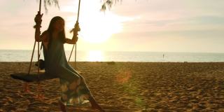 亚洲美女坐在海边的秋千上与夕阳背景在周末假期。快乐独自旅行的概念。4 k慢动作。