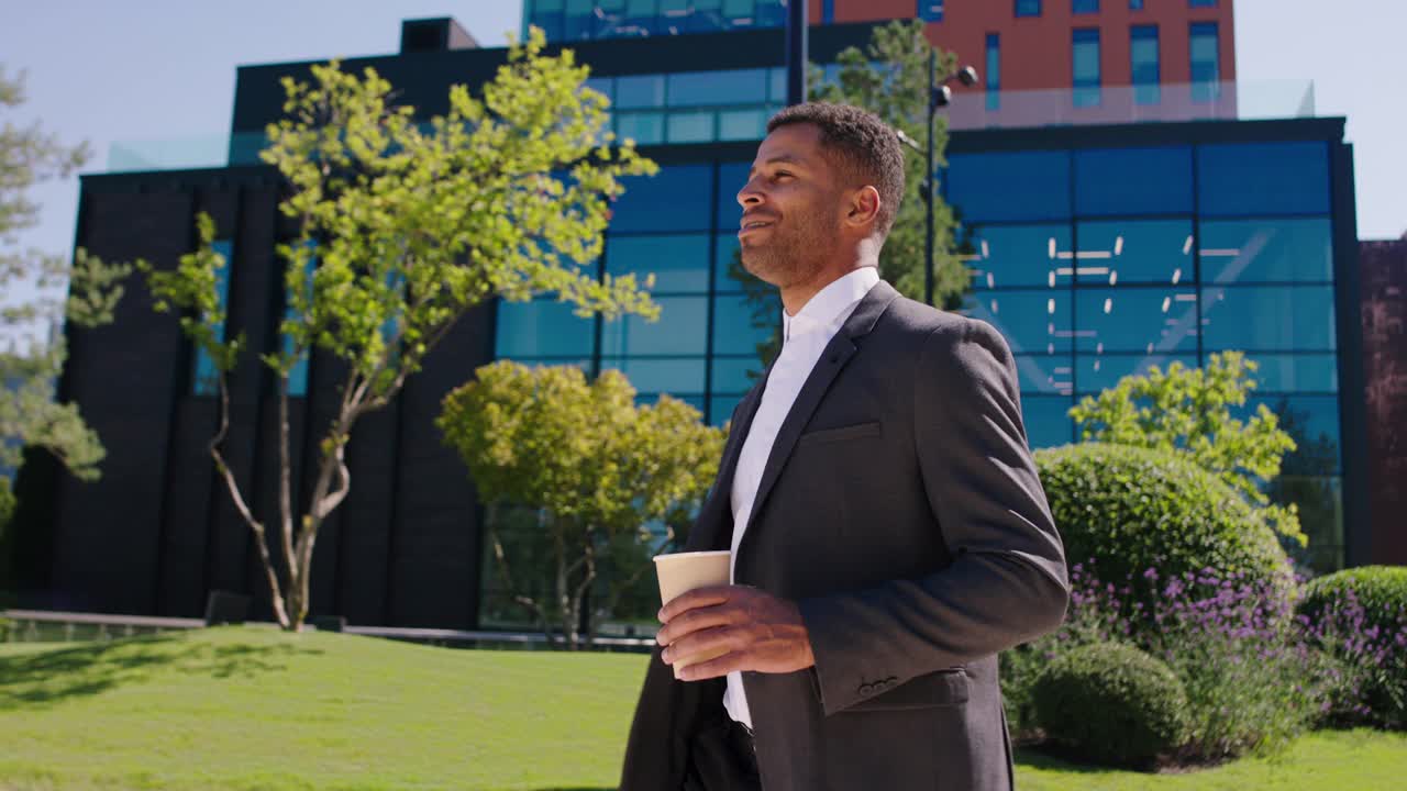 有魅力的办公室经理黑人男人愉快地走在现代商业中心大楼旁边他拿着一杯咖啡