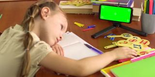 一个小女孩在桌子边睡觉，手里拿着一部绿色屏幕的智能手机。远程学习。