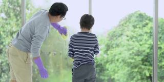 肖像的亚洲父亲和小儿子在橡胶手套与洗涤剂和抹布清洗窗户玻璃一起。男孩帮爸爸打扫房子。日常工作的概念。