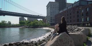 纽约，一名身穿黑衣的女子坐在水边的石头上。伸展手臂了