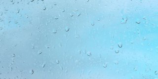水滴顺着透明的玻璃流下来，雨滴落在窗户上，形成液体的气泡，在背景中是美丽的蓝天。