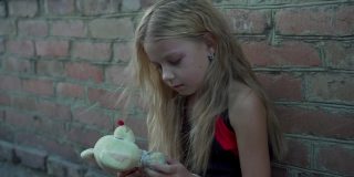 一个来自贫困地区的悲伤小女孩靠着一堵砖墙坐在那里，手里拿着一个旧的填充玩具。