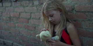一个来自贫困地区的悲伤小女孩靠着一堵砖墙坐在那里，手里拿着一个旧的填充玩具。