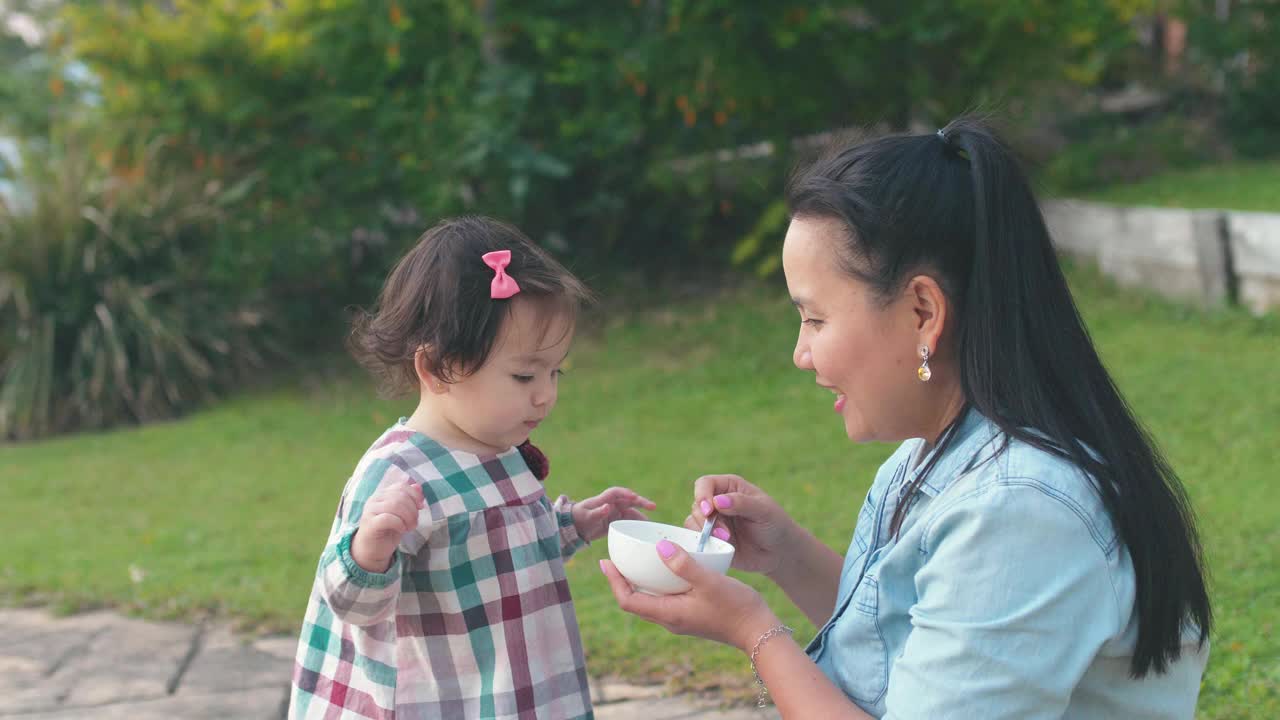 亚洲母亲在前院喂养她的混合蹒跚学步的女儿，家庭生活理念