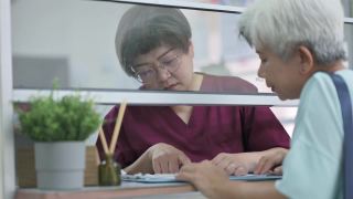 亚裔华人老年女性在诊所用信用卡支付医疗费用，由女护士手持信用卡读卡器视频素材模板下载