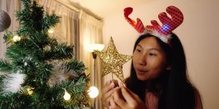快乐的亚洲女孩戴着驯鹿发带，晚上在家装饰圣诞树，庆祝节日的概念。