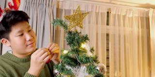 快乐的亚洲男孩戴着驯鹿发带，晚上在家装饰圣诞树，庆祝节日的概念。