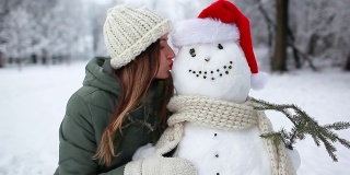 在下雪的冬天公园里，女人戴着圣诞帽和围巾亲吻雪人。圣诞节的庆祝活动。户外活动